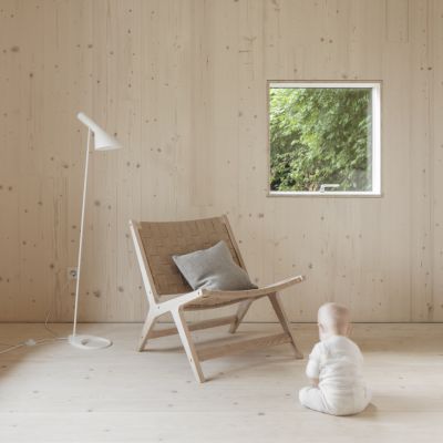 Child in a white onesie sits on a light wooden floor in front of a wooden chair with a grey cushion, next to a white floor lamp and a window with a view of greenery.